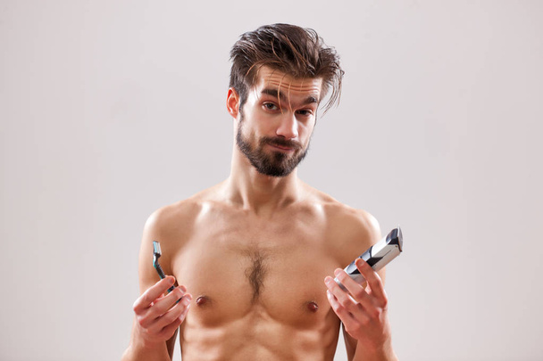 Νεαρός άνδρας κρατά ηλεκτρικών και κλασικό ξυράφι. Αυτός να αποφασίσει ποιο να χρησιμοποιήσετε για να ξυρίσει τα γένια του.  - Φωτογραφία, εικόνα