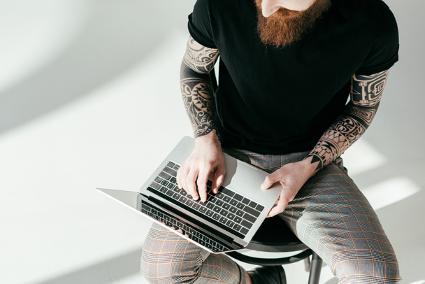 cropped image of bearded tattooed man sitting on chair with laptop on white - Zdjęcie, obraz