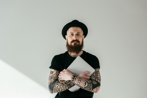 handsome bearded tattooed man hugging laptop and looking at camera on white - Photo, Image