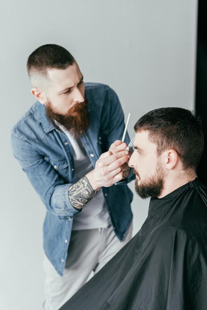 vista lateral del peluquero cortando el pelo del cliente en la barbería aislado en blanco
 - Foto, imagen