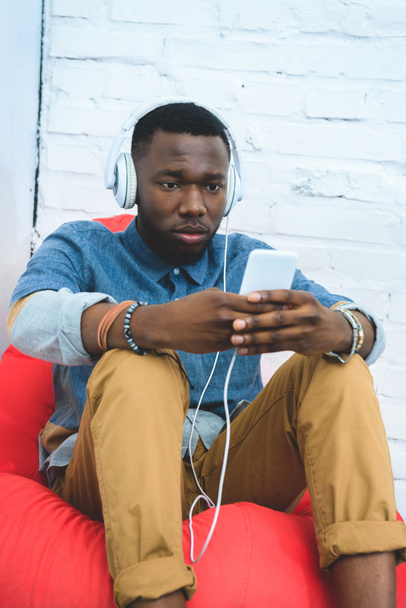 Przystojny mężczyzna Afroamerykanów w słuchawki do słuchania muzyki na smartphone, siedząc w worek fasoli - Zdjęcie, obraz