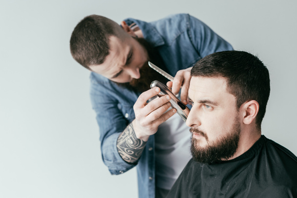 serious barber trimming customer beard at barbershop isolated on white - Fotó, kép