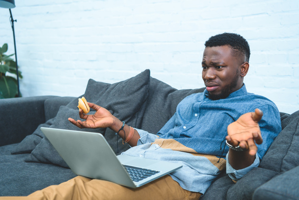 verwirrter afrikanisch-amerikanischer Mann arbeitet am Laptop, während er auf dem Sofa sitzt und Hamburger isst - Foto, Bild