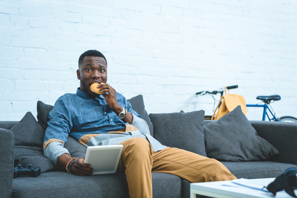 Handsome african american man using tablet and eating hamburger - 写真・画像