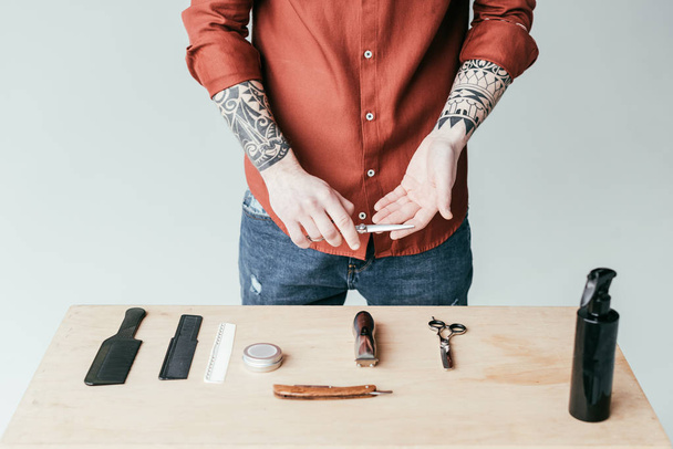 cropped image of tattooed barber holding scissors above table with equipment isolated on white - Valokuva, kuva