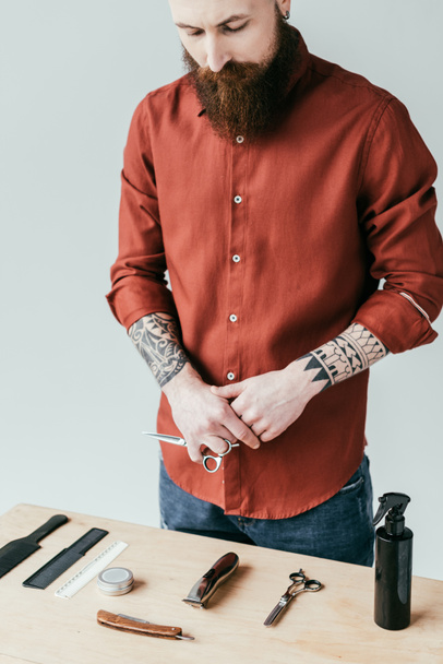 bärtiger, gutaussehender Friseur, der eine Schere in der Hand hält und die Ausrüstung isoliert auf weißem Grund betrachtet - Foto, Bild