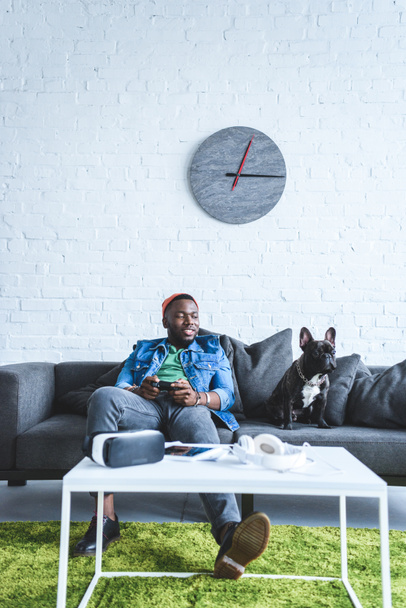 junger Mann mit Steuerknüppel auf dem Sofa sitzend mit Bulldogge und digitalen Geräten auf dem Tisch - Foto, Bild