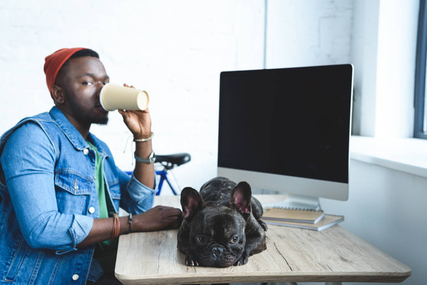 Young man drinking coffee by French bulldog lying on computer table - Фото, зображення