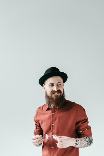 smiling bearded man in hat holding glasses isolated on white - Фото, изображение