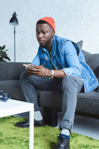 African american man sitting on sofa and using smartphone - Фото, изображение