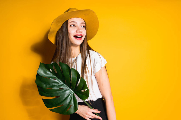 stylish smiling girl with long hair wearing a trendy hat, holding a green leaf and posing on an orange background - Foto, imagen