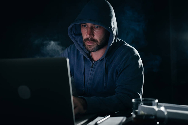 criminal man with a hood on his head hacks a laptop in the dark - Photo, Image