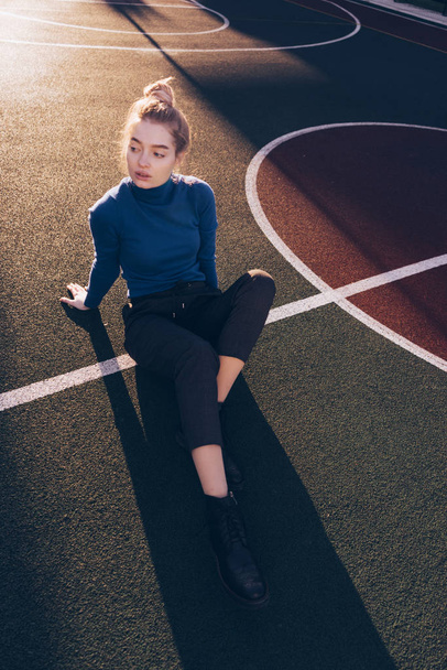 model Slim mladá dívka v modrém svetru sedí na sportovní hřiště venku, v paprscích ranního slunce - Fotografie, Obrázek