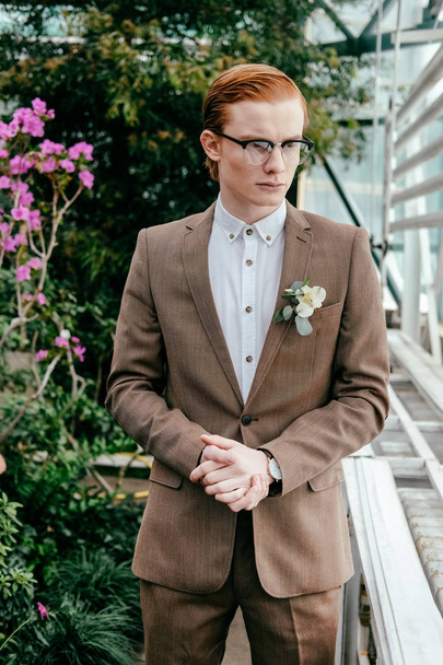 portrait of stylish man with red hair in suit and eyeglasses looking away in greenhouse - Fotó, kép