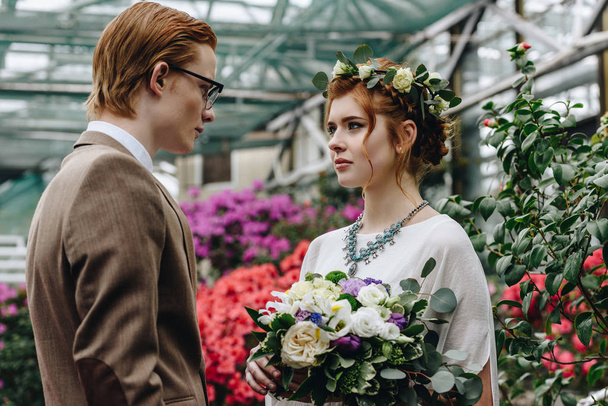 stylish young groom and beautiful redhead bride looking at each other in botanical garden - Fotoğraf, Görsel