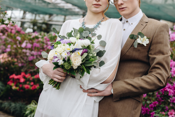 スタイリッシュな若い新郎と植物園で一緒にウェディング ブーケの立っている花嫁のショットをトリミング - 写真・画像