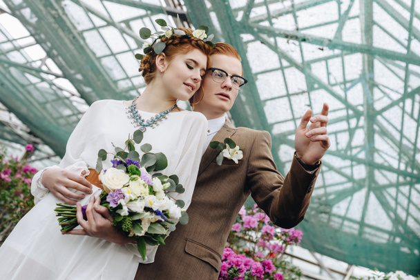 low angle view of beautiful young redhead wedding couple standing together and looking away in botanical garden - Foto, imagen