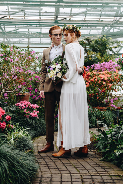 vista completa de hermosa pareja de boda elegante joven de pie juntos entre las flores en el jardín botánico
 - Foto, Imagen