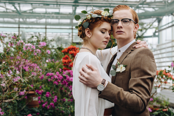 tender young red-haired wedding couple embracing while standing between flowers in botanical garden - 写真・画像