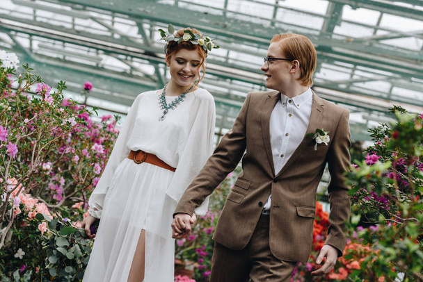 Szczęśliwa młoda redhead ślub para trzymając się za ręce i chodzenia między kwiaty w ogrodzie botanicznym - Zdjęcie, obraz