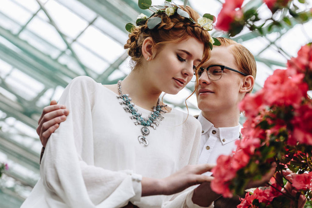 glücklicher junger Bräutigam mit Brille, der die schöne rothaarige Braut im Blumenkranz betrachtet - Foto, Bild