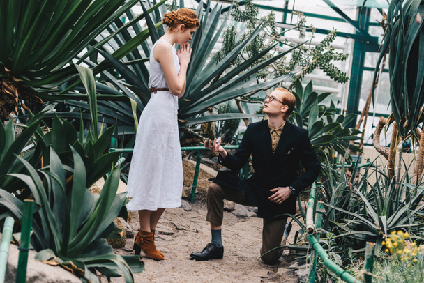 stylish young man standing on knee and making proposal to beautiful red-haired girl in botanical garden  - Photo, Image