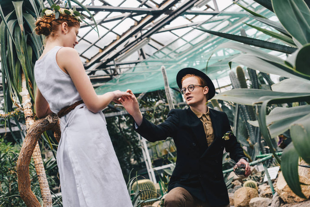 joven con estilo en sombrero y anteojos haciendo propuesta a hermosa chica en el jardín botánico
 - Foto, imagen