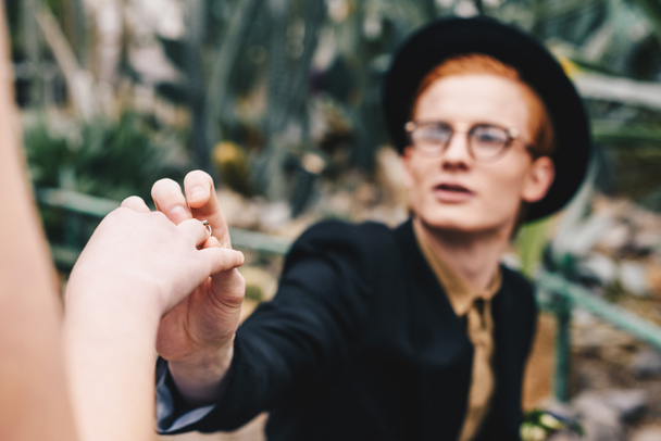 tiro cortado de jovem elegante em chapéu e óculos fazendo proposta e colocando anel para menina
 - Foto, Imagem