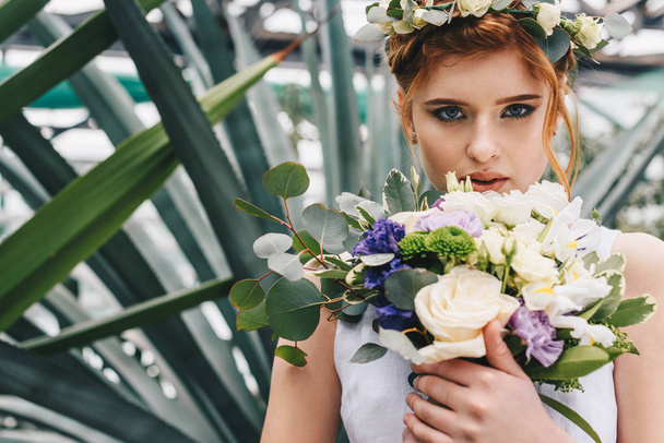 beautiful young red-haired bride holding wedding bouquet and looking at camera  - Foto, imagen