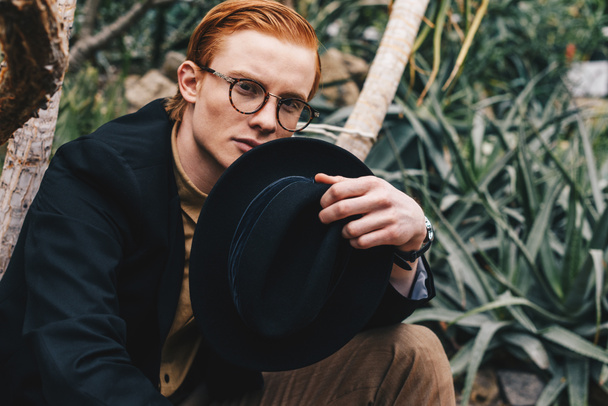 csinos fiatal vörös hajú férfi szemüvegek gazdaság kalap és látszó-on fényképezőgép - Fotó, kép