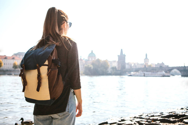 A beautiful young tourist girl with a backpack stands next to the Vltava river in Prague and admires one of the most popular attractions called the Charles Bridge. Travel around the Czech Republic. - Zdjęcie, obraz