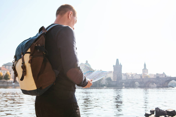 A young tourist man with a backpack standing next to the river Vltava in Prague looks at the map and admires the architecture of the city. Charles Bridge is near. Travel around the Czech Republic. - Photo, Image
