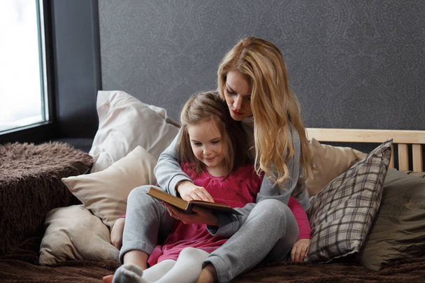 Mother and daughter sitting on the bed in an embrace and reading a book - Photo, image
