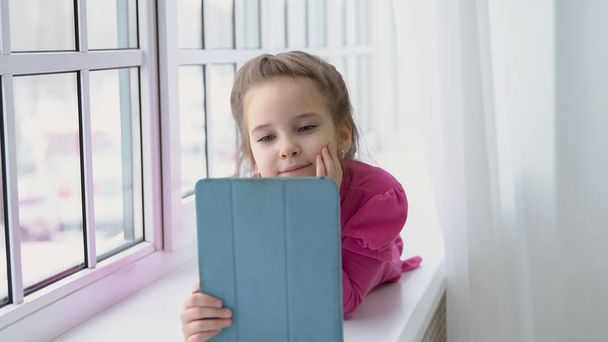Μικρό κορίτσι σε ένα ροζ φόρεμα κάθεται δίπλα στο παράθυρο με ένα tablet - Φωτογραφία, εικόνα