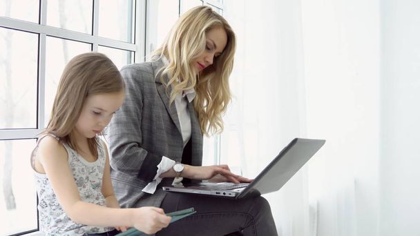 Young business woman sitting by the window with her daughter and laptop - Foto, Imagem
