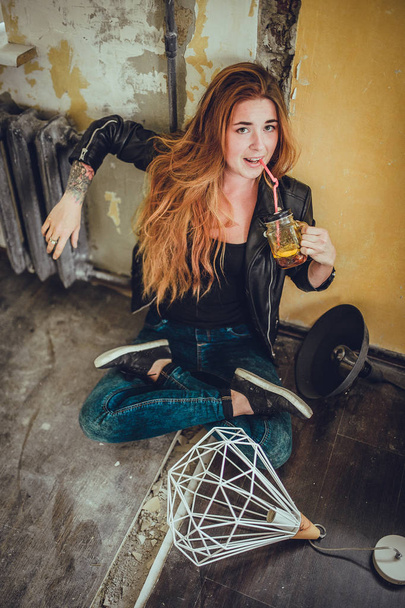 portrait of young fashion woman with red hair and tattoo in black clothes sitting on floor at vintage style room. drink fresh cocktail  - Φωτογραφία, εικόνα