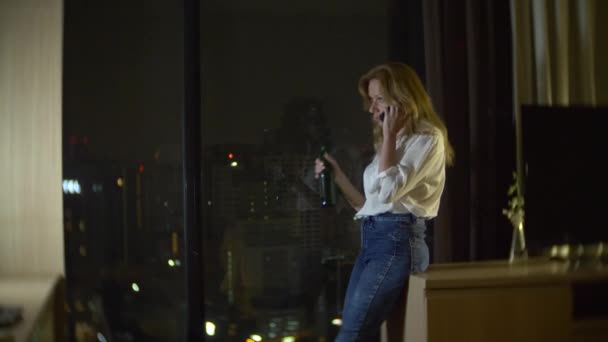 a woman is drinking beer at home next to a panoramic window overlooking the skyscrapers. night, background blur. 4k, slow motion - Felvétel, videó