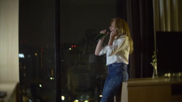 a woman is drinking beer at home next to a panoramic window overlooking the skyscrapers. night, background blur. 4k, slow motion - Felvétel, videó