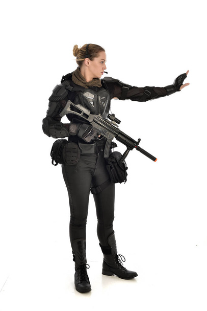 戦術的な黒の鎧を着て、白いスタジオ背景に分離されたライフル銃を保持している女性兵士の完全な長さの肖像画. - 写真・画像