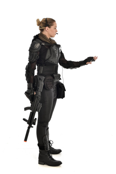戦術的な黒の鎧を着て、白いスタジオ背景に分離されたライフル銃を保持している女性兵士の完全な長さの肖像画. - 写真・画像