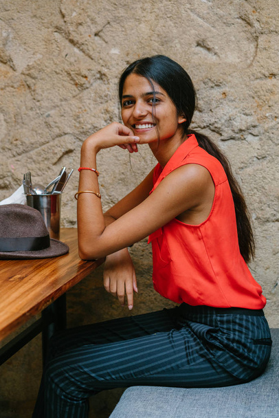 Portrait of a young and attractive Indian Asian woman smiling at the camera. She is seated and has a brown fedora cap . She is dressed in smart casual and is relaxed and cheerful. - Fotografie, Obrázek