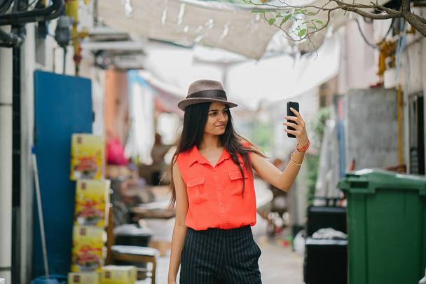  young and stylish Indian Asian woman takes a selfie of herself in an alley way in Asia. She is smiling and posing for the shot in the sun. - Foto, Imagem