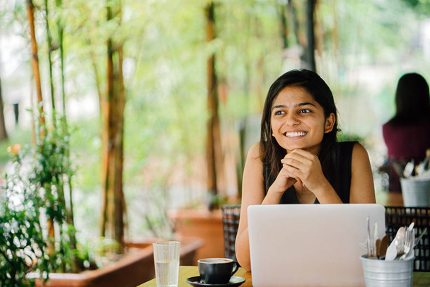 Candid portrait of an attractive and young Indian Asian professional woman work on her laptop in a trendy cafe or coworking space with teal wooden furniture. She is smiling and looking away. - Fotoğraf, Görsel