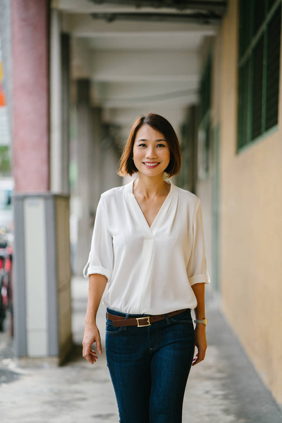  young Chinese Asian woman walks along a corridor in Singapore, Asia. She is dressed smartly in business casual, has bobbed hair and is smiling as she strolls down the corridor. - Zdjęcie, obraz