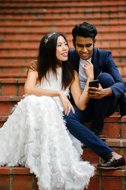  interracial couple are engaged and sit on brick steps and are looking at a smartphone together. They are smiling. The Chinese woman is in an elegant white wedding dress and the man in a smart suit. - Photo, image