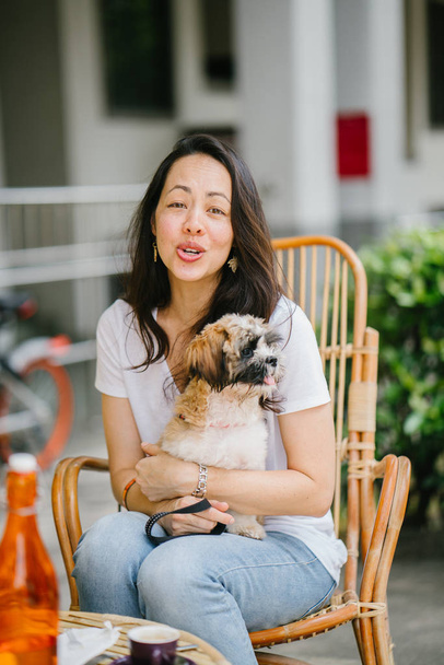  young Pan Asian woman enjoys her weekend with her pet shih tzuh dog. She is sitting in a rattan chair in the sun in a cafe with her dog  - Фото, изображение