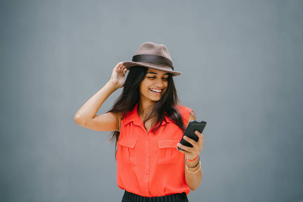 Portrait of a young and fashionable North Indian Desi Asian woman. She has a light complexion and is wearing an orange shirt, black pants and a fedora hat. She smiles as she uses her smartphone. - Fotó, kép