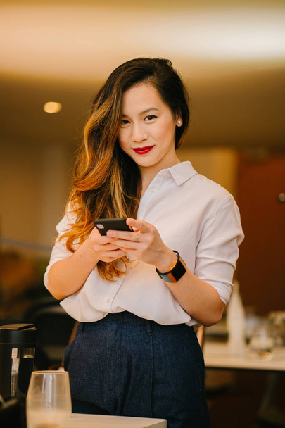 Portrait of a young and attractive Chinese Asian woman smiling as she uses her phone. She is very professionally dressed and is in an office or coworking space. - Фото, зображення