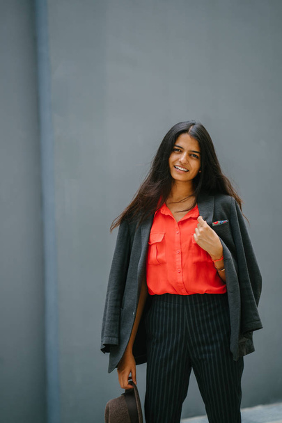 Portrait of a fashionable Indian Asian woman against a plain grey background. She is smiling and wearing a smart orange blouse and black pinstripe pants with a jacket draped over her shoulders. - Valokuva, kuva