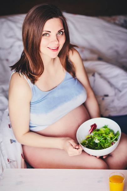 Pregnant smiling girl in T-shirt sit on bed and eat salad with green and red leaves, drink orange, pumpkin and carrot juice. Young woman keep diet during pregnancy. Healthy food for expectant mothers. - 写真・画像